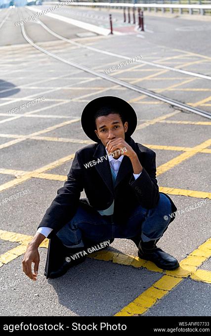 Stylish young man wearing hat crouching on street during sunny day