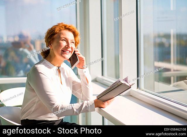 Happy confident businesswoman talking over mobile or smart phone and looking at window. Red haired lady communicating with foreign partners and holding notebook...