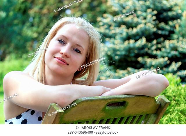 Closeup portrait of cool smiling young blond woman sitting on the wooden chair in the garden. The woman has a slightly bowed head and is looking at the camera
