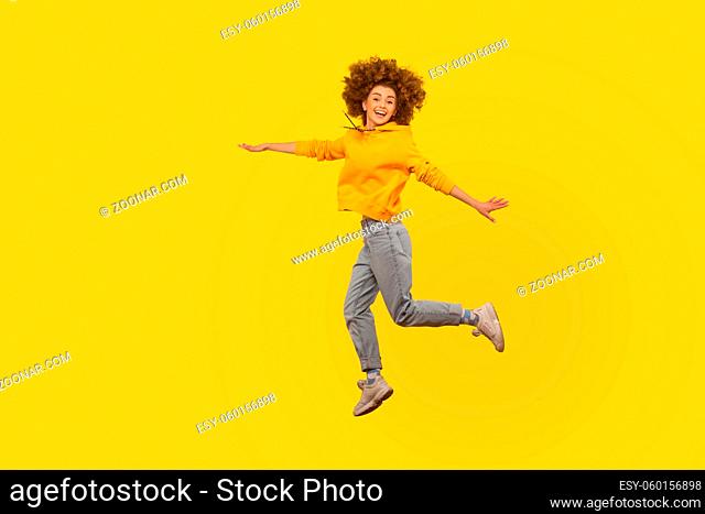 Portrait of enthusiastic lively energetic curly-haired girl in urban style hoodie and jeans flying walking in air, enjoying happy life, feeling inspired