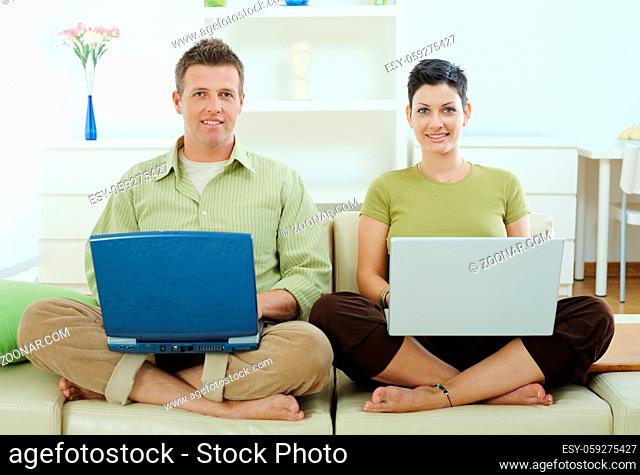 Happy young couple sitting on couch at home using laptop computer, smiling