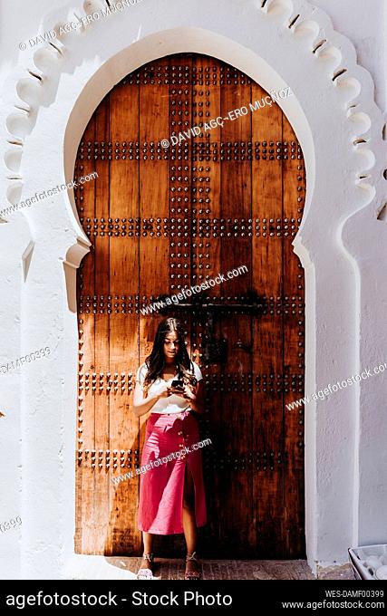 Young woman standing in front of traditional entrance door looking at cell phone, Merzouga, Morocco