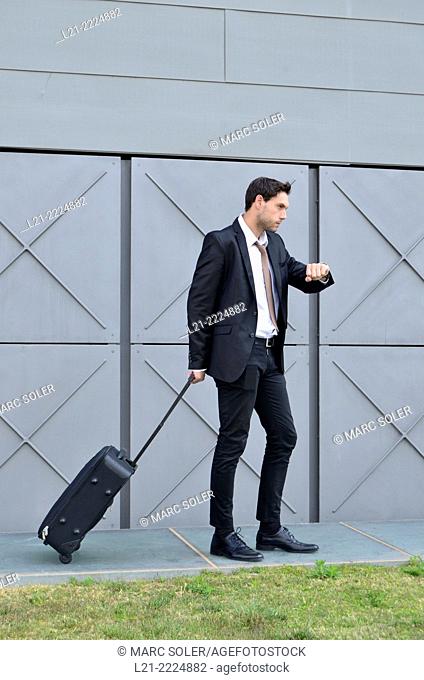 Businessman with wheeled suitcase looking at wrist watch