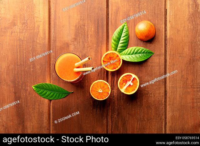 A glass of fresh orange juice with blood oranges, green leaves, and two bamboo straws, shot from above on a dark rustic wooden background with a place for text