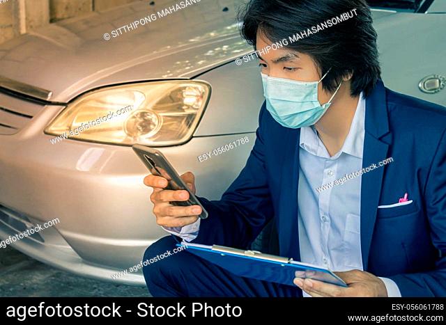 Sitting Asian Insurance Agent or Insurance Agency in Suit Wear Mask and See Smartphone and Hold Report Inspecting Car from Accident for Claim at Garage in...