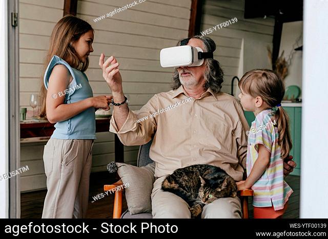 Grandfather wearing virtual reality simulator gesturing by grandchildren at home