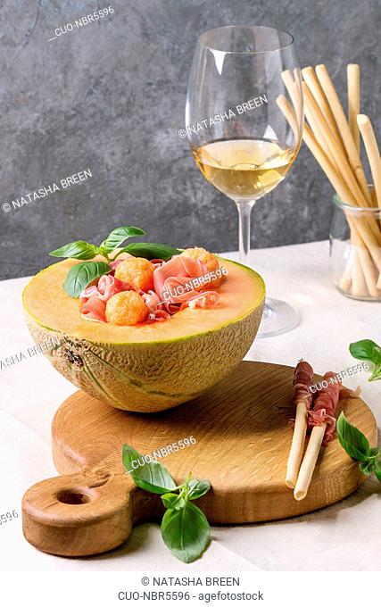 Melon and ham or prosciutto salad served in half of Cantaloupe melon, decorated by fresh basil standing on wooden serving board over white tablecloth with glass...