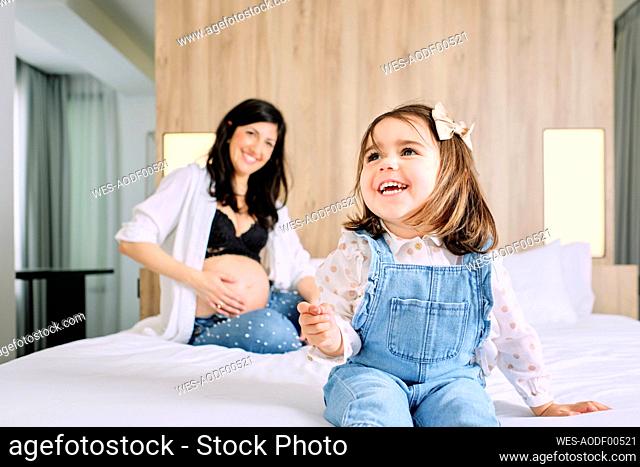Cheerful daughter and pregnant mother sitting on bed in bedroom