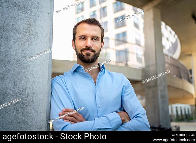 Smiling businessman with arms crossed leaning on wall in front of building