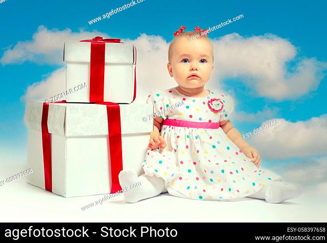 Adorable baby girl sit on gift box point finger aside posing over cloudy sky background. New Year and Christmas celebration concept