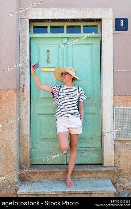 Beautiful young female tourist woman wearing big straw hat, taking self portrait selfie, standing in front of turquoise vinatage wooden door and textured stone...