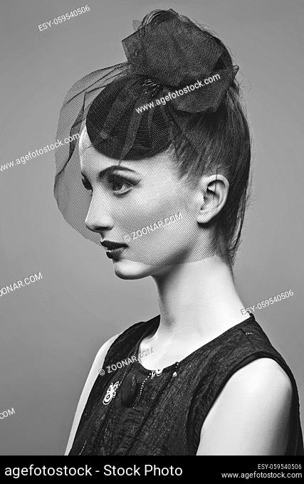 Beautiful young woman in small black veil hat cap with hairdo and bright make-up. Doll style. Beauty shot on grey background. Copy space. Monochrome