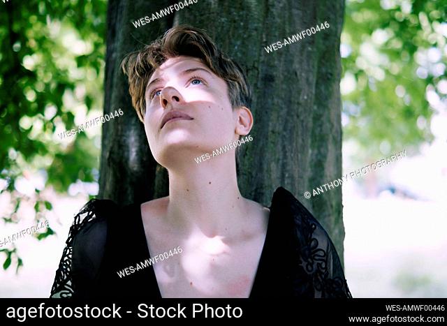 Thoughtful non-binary person leaning on tree trunk