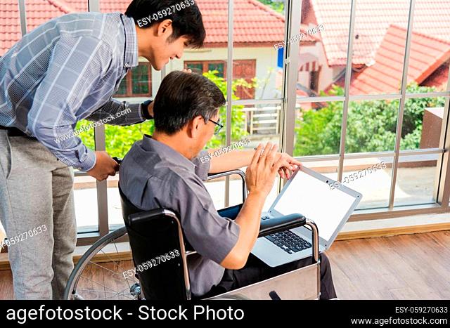 Asian senior disabled businessman in a wheelchair with laptop computer discuss together with team in office. Old father man sitting wheelchair and his son...