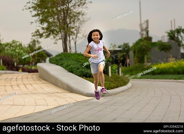 Young girl with long hair running down an outdoor park path and looking at the camera; Hong Kong, China