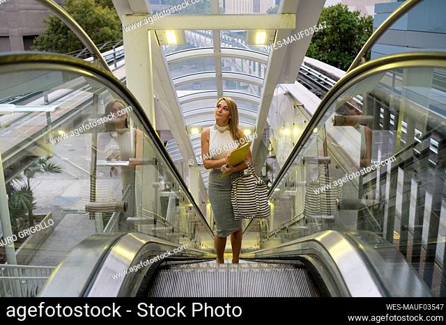 Thoughtful blond female professional looking up while standing on escalator in city