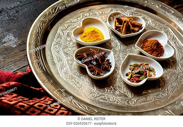 Five heart shaped trays with star anise and curry powder besides other spices to form a flower on a round silver platter