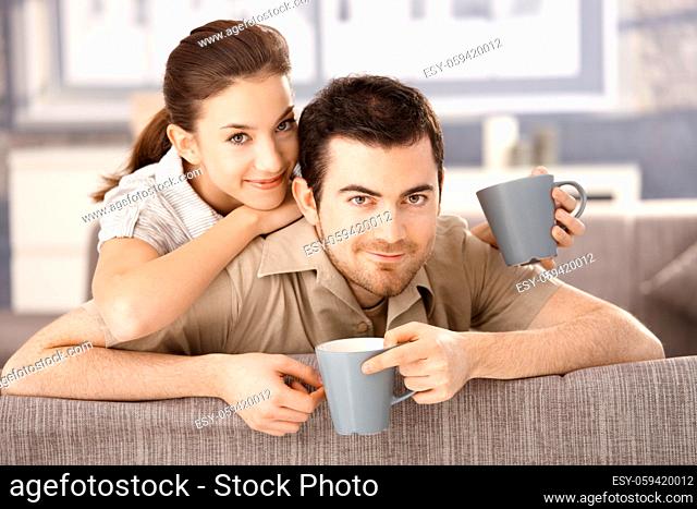 Happy couple sitting on sofa at home, drinking tea, smiling