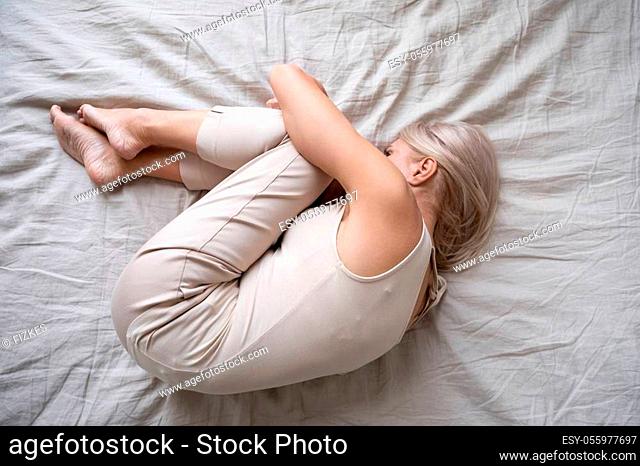 View from top depressed middle-aged alone female curled up in fetal position lying in bed, concept of chronic insomnia, sleeping disorder