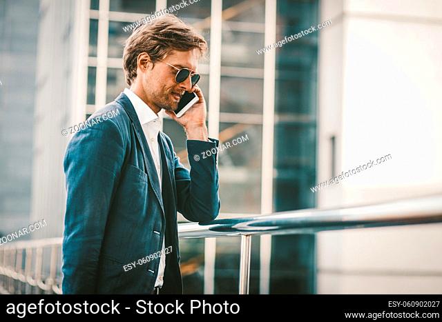 Handsome man walking on a bridge and talking on the phone. Happy smiling young businessman standing outside and having conversation on the phone with urban...