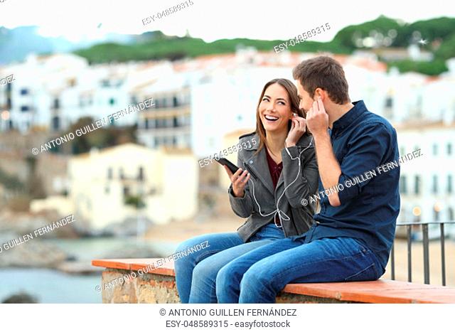 Happy couple sharing online music from a smart phone sitting on a ledge on vacation in a coast town
