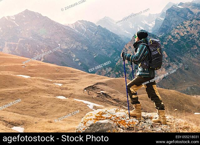 A guy with a beard and wearing sunglasses in a membrane jacket, hat, with a backpack and sticks for Nordic walking, a traveler standing in the open air and...