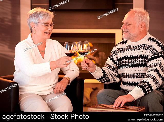 Retired husband and wife drinking red wine together in cosy living room, in front of fireplace, looking happy, smiling