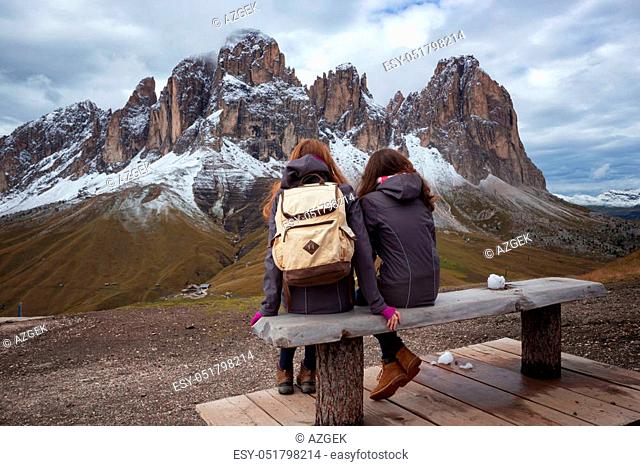 Two girls are sitting on a bench and view of Sassolungo Langkofel. Dolomites, Italy