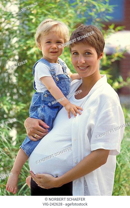 portrait, half-figure, outdoor, pregnant woman with short brown hair wearing a white shirt stands with her 2-year-old blond son on her arm in the garden  -...