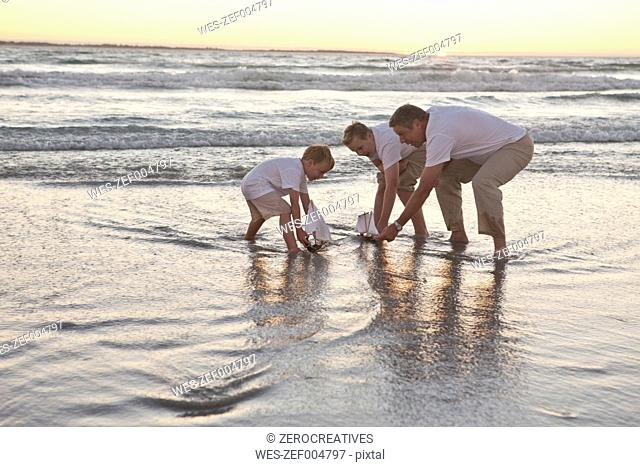 Father and sons playing with toy wooden boats in the water on a beach