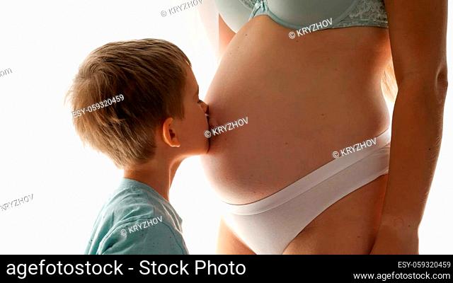 Closeup of little boy kissing his pregnatn mom in big belly standing at window in bedroom. Concept of family happiness and baby anticipation