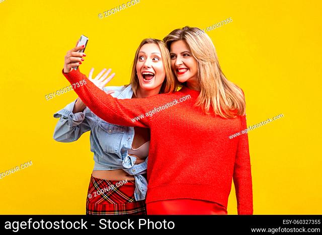 Best friends having fun. Portrait of happy women in stylish clothes standing with toothy smile and taking selfie on phone together, enjoying life