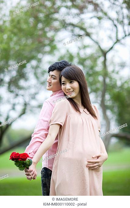 Pregnant woman and young man standing with back to back and holding flowers with smile