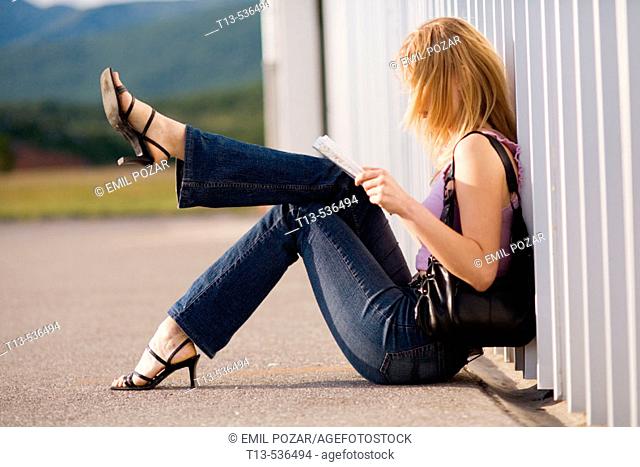 Young blonde woman is reading a road map while sitting by the wall with one leg lifted hight into air