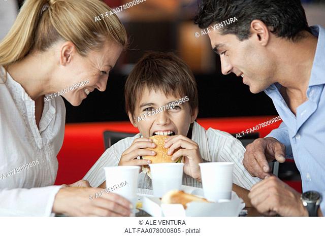 Boy eating hamburger with his parents in fast food restaurant