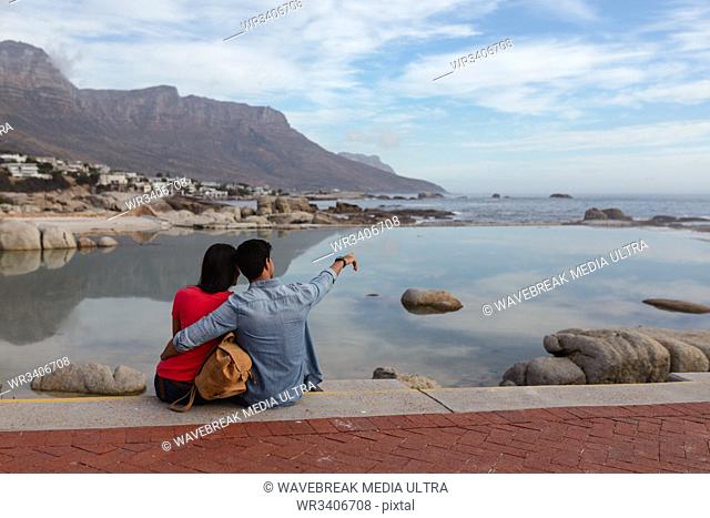 Rear view of a young mixed race couple sitting outside on a wall embracing and looking out to sea, the man is pointing to the distance