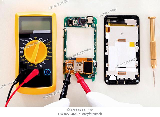 High Angle View Of Person Checking Damaged Mobile Phone With Digital Multimeter
