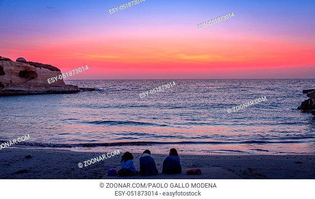 Three women waiting for the sunrise in front of the sea (Puglia region, South of Italy). Concept of frienship, travel and adventure