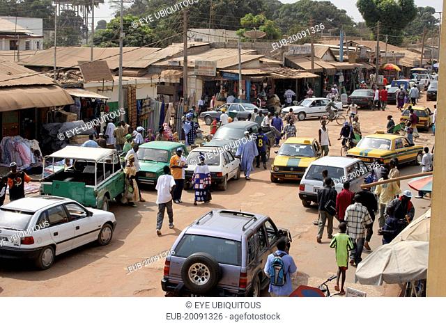 Bakau Market, Atlantic Road. View of Serekunda's main city street lined with open fronted shops and roadside stalls beneath sun umbrellas, people and vehicles