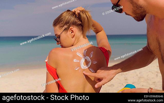 Happy couple applying sun tanning lotion on the beach, man putting sunscreen suntan cream on woman