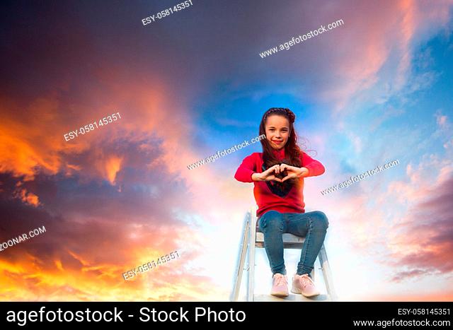 The teenage girl is sitting on top of a metal ladder. The girl sits on a background of colored sky and shows heart with her fingers