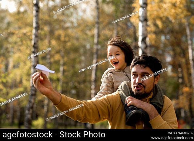 Playful man playing with paper airplane carrying son on shoulders in park