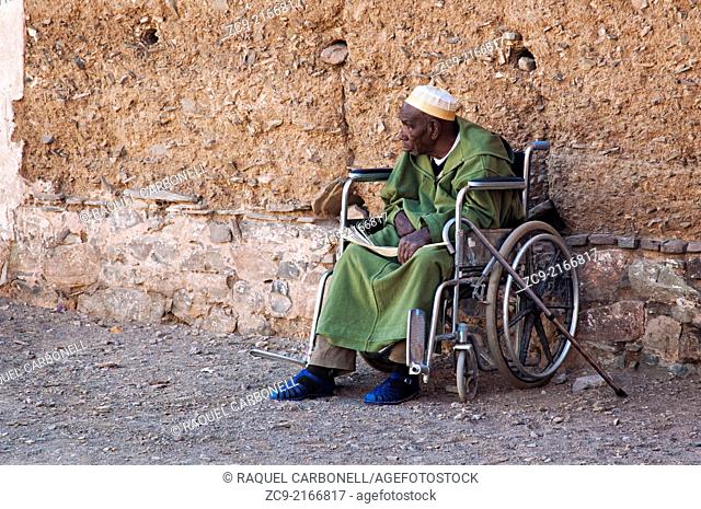 Portrait of an elder man in djellaba sitting on a wheelchair on the streets of a traditional sdobe village, route between Foum Zguid and Tata, Morocco