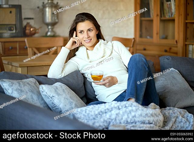 attractive young woman in sweater relaxes on a gray sofa at home and drinks tea with lemon
