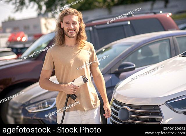 Good mood. Young adult friendly man standing near car at gas station smiling at camera on sunny day