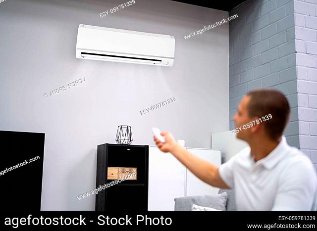 Young Happy Man Sitting On Couch Operating Air Conditioner With Remote Control At Home