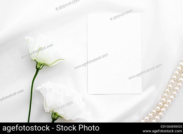 Wedding invitation, white rose flowers and pearls on silk fabric as bridal flatlay background, blank paper greeting card and holiday branding