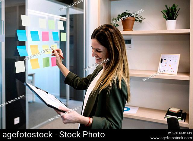 Smiling mid adult businesswoman holding clipboard while planning strategy on adhesive note at office