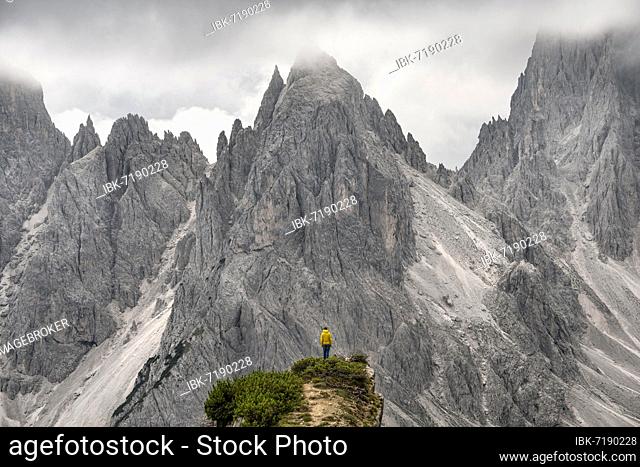 Hiker standing on a ridge, mountain peaks and pointed rocks behind, dramatic cloudy sky, Cimon di Croda Liscia and Cadini group, Auronzo di Cadore, Belluno