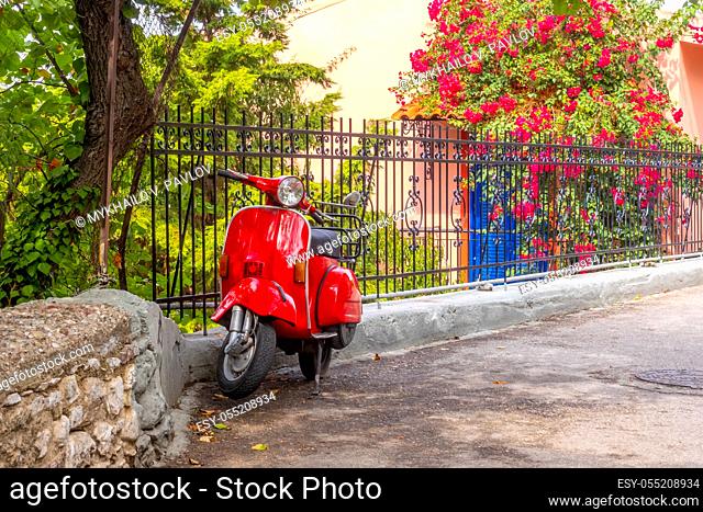 Summer blooming garden in sunny weather. A red scooter in retro style is parked at the fence
